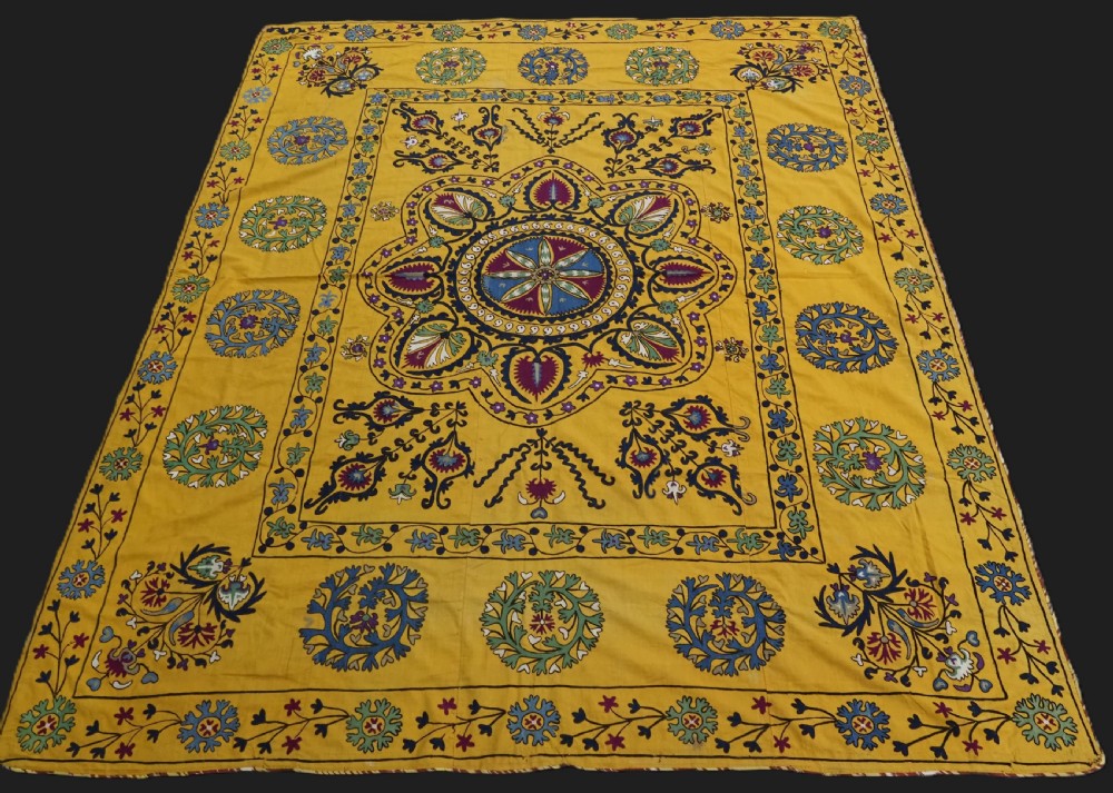 A rectangular yellow cotton textile featuring hand-embroidered silk floral medallions, vines, and circular motifs in blue, green, and burgundy.