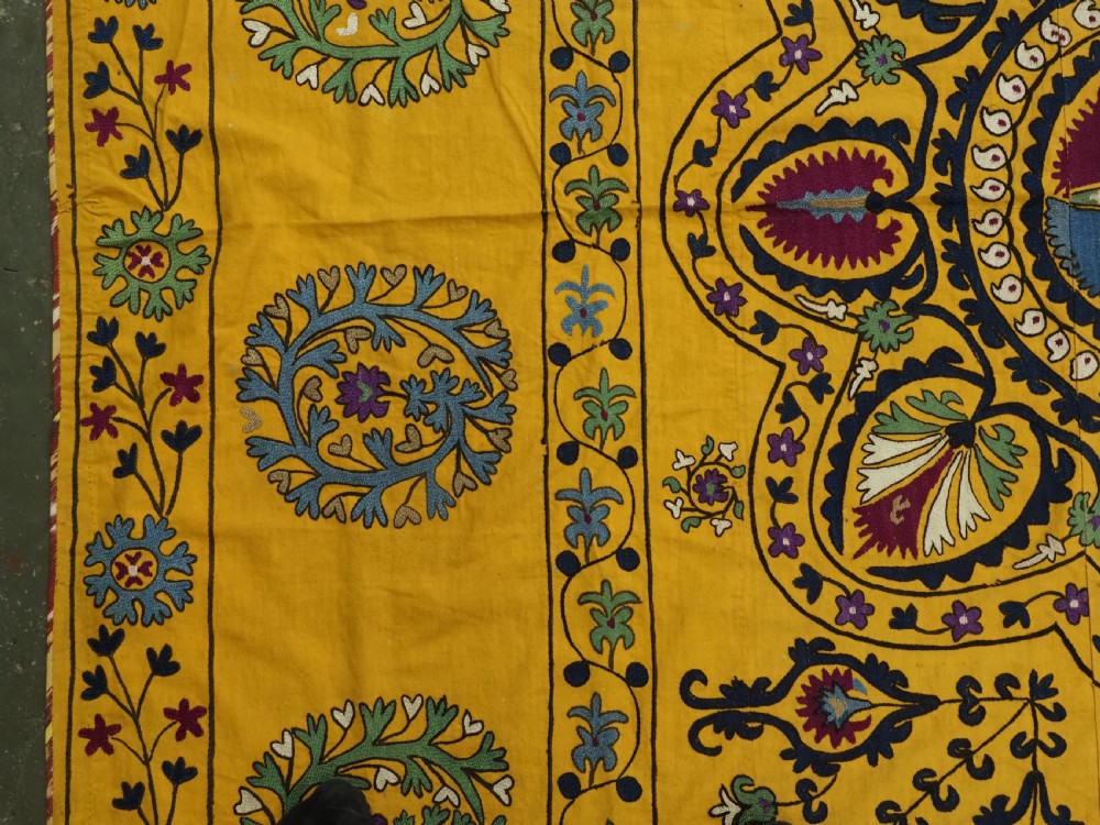 A rectangular yellow cotton textile featuring hand-embroidered silk floral medallions, vines, and circular motifs in blue, green, and burgundy. View III