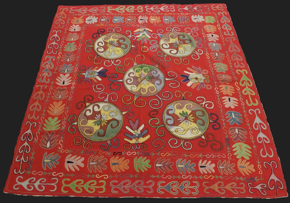 A rectangular red wool textile featuring multi-coloured silk embroidery with central circular medallions and decorative botanical borders.