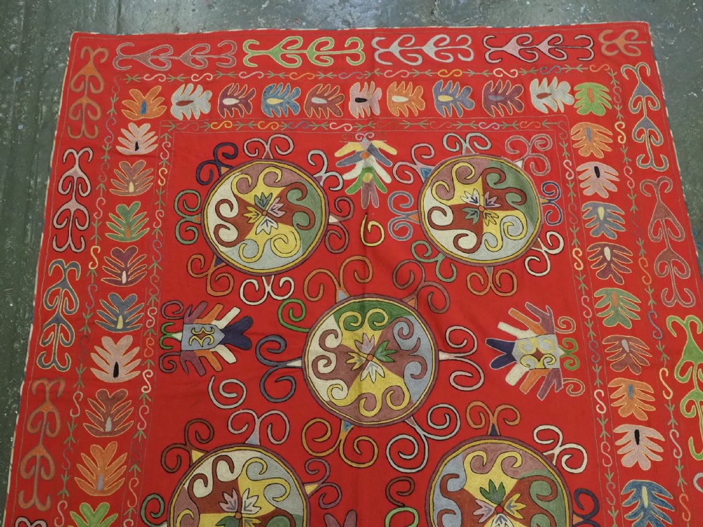 A rectangular red wool textile featuring multi-coloured silk embroidery with central circular medallions and decorative botanical borders. View II