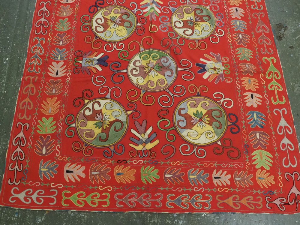A rectangular red wool textile featuring multi-coloured silk embroidery with central circular medallions and decorative botanical borders. View III