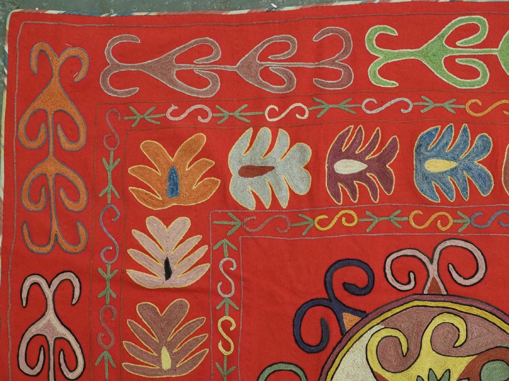 A rectangular red wool textile featuring multi-coloured silk embroidery with central circular medallions and decorative botanical borders. View IV