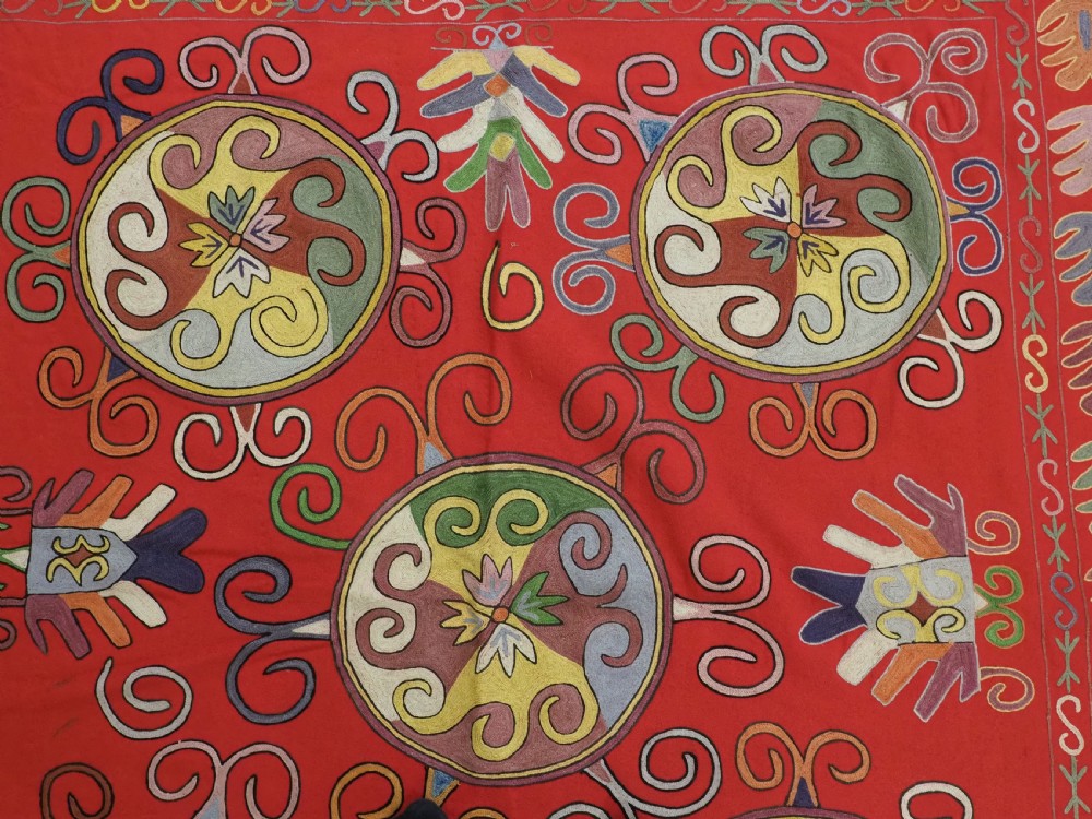 A rectangular red wool textile featuring multi-coloured silk embroidery with central circular medallions and decorative botanical borders. View VI
