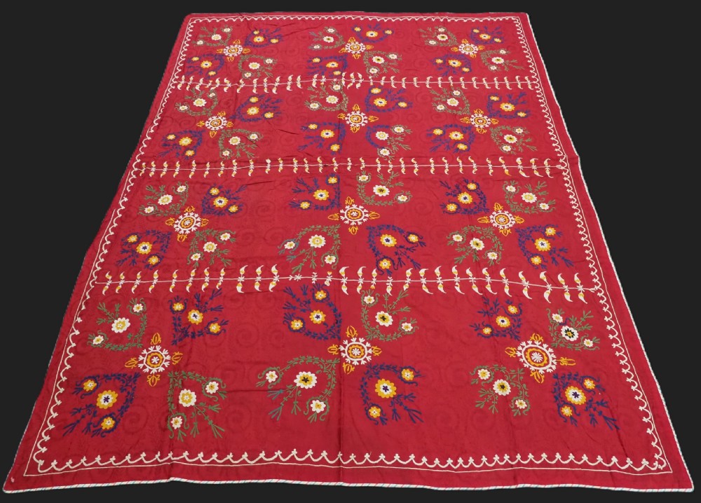 An antique rectangular red cotton suzani textile featuring hand embroidery in silk with floral rosettes and botanical motifs in white, yellow, green, and blue.