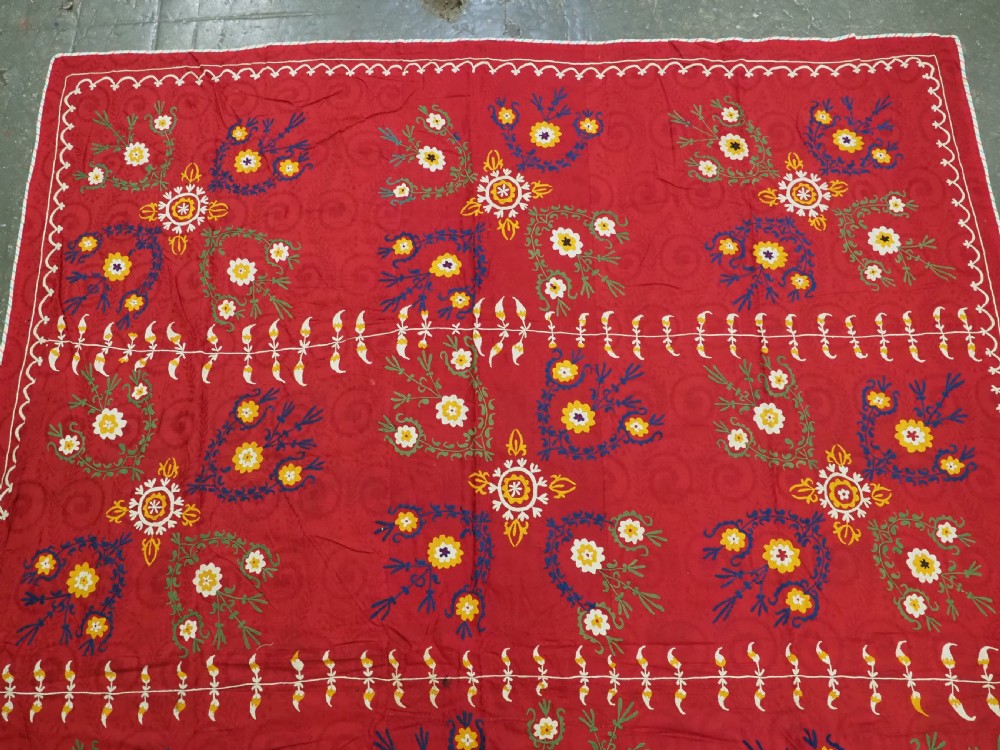 An antique rectangular red cotton suzani textile featuring hand embroidery in silk with floral rosettes and botanical motifs in white, yellow, green, and blue. View II