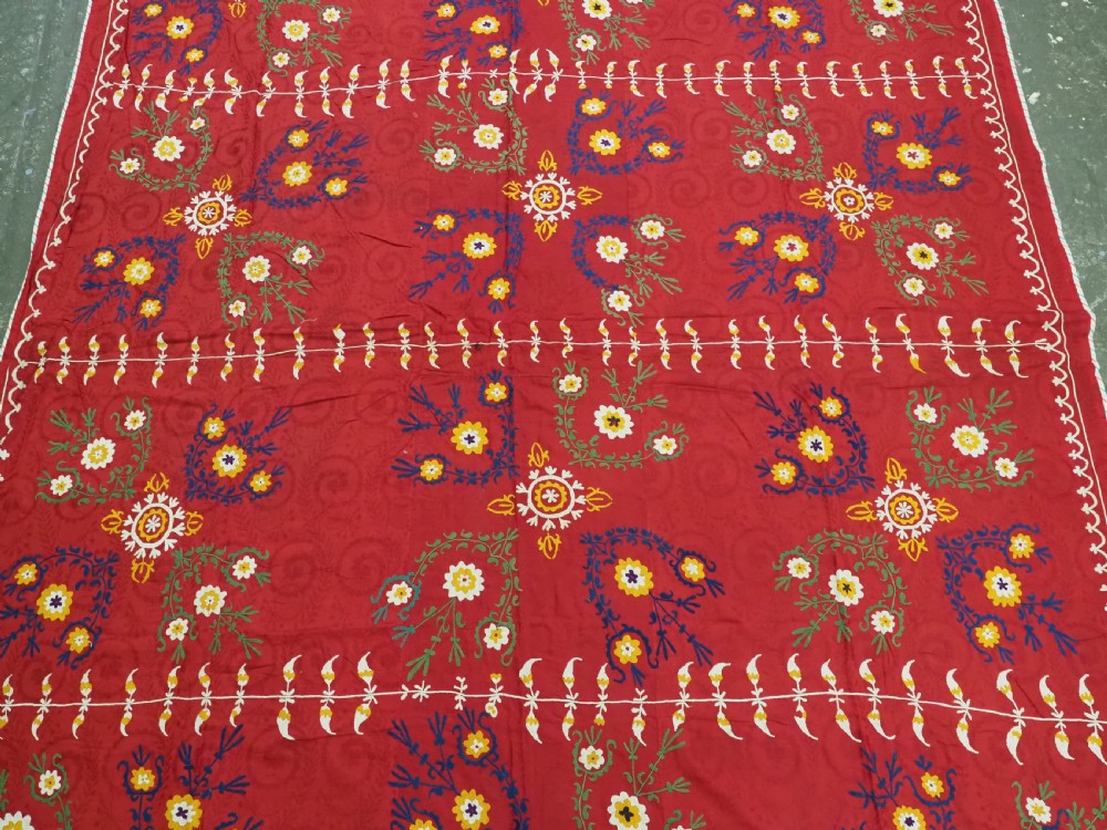 An antique rectangular red cotton suzani textile featuring hand embroidery in silk with floral rosettes and botanical motifs in white, yellow, green, and blue. View III