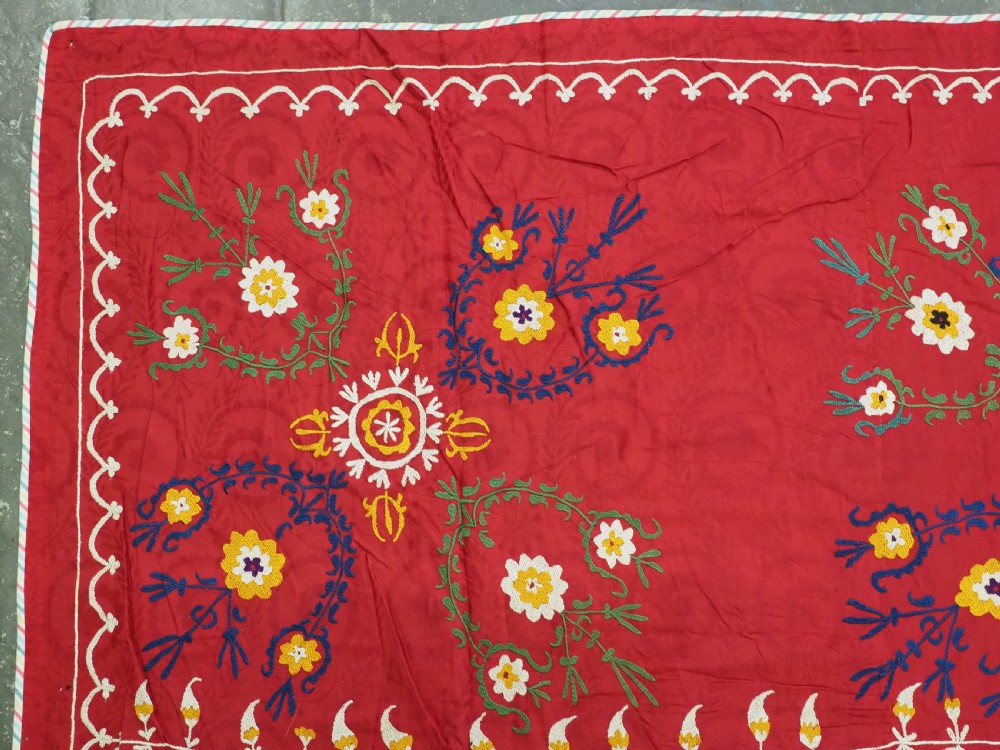 An antique rectangular red cotton suzani textile featuring hand embroidery in silk with floral rosettes and botanical motifs in white, yellow, green, and blue. View V