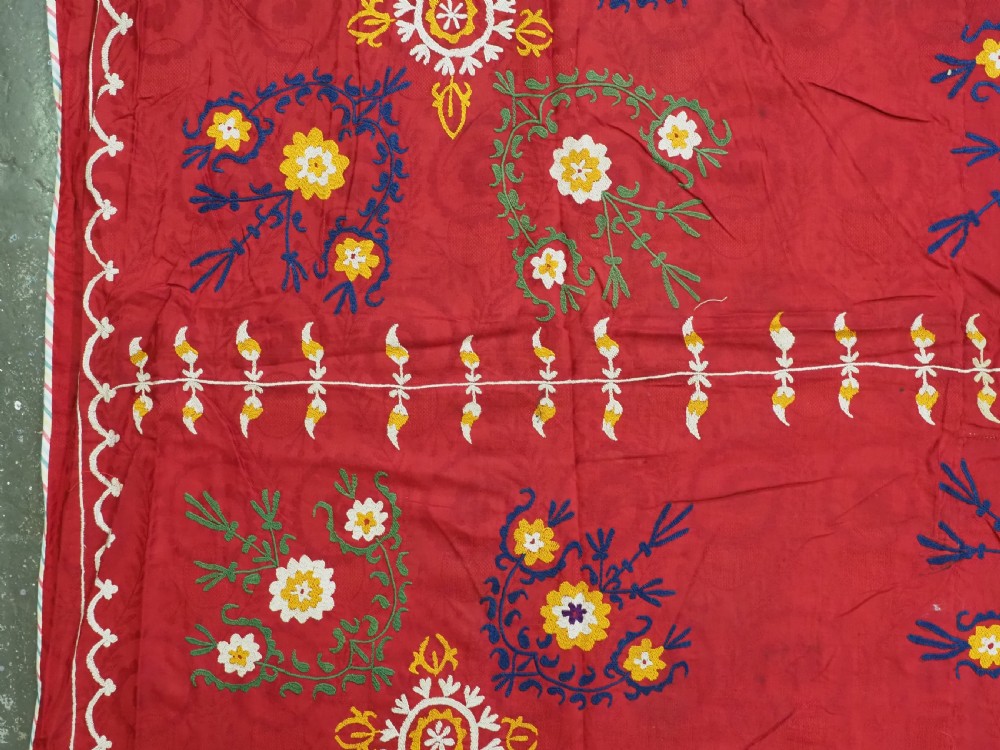 An antique rectangular red cotton suzani textile featuring hand embroidery in silk with floral rosettes and botanical motifs in white, yellow, green, and blue. View VII