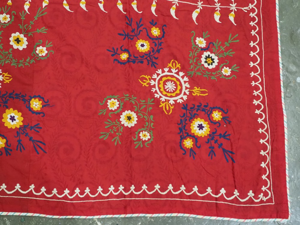 An antique rectangular red cotton suzani textile featuring hand embroidery in silk with floral rosettes and botanical motifs in white, yellow, green, and blue. View X