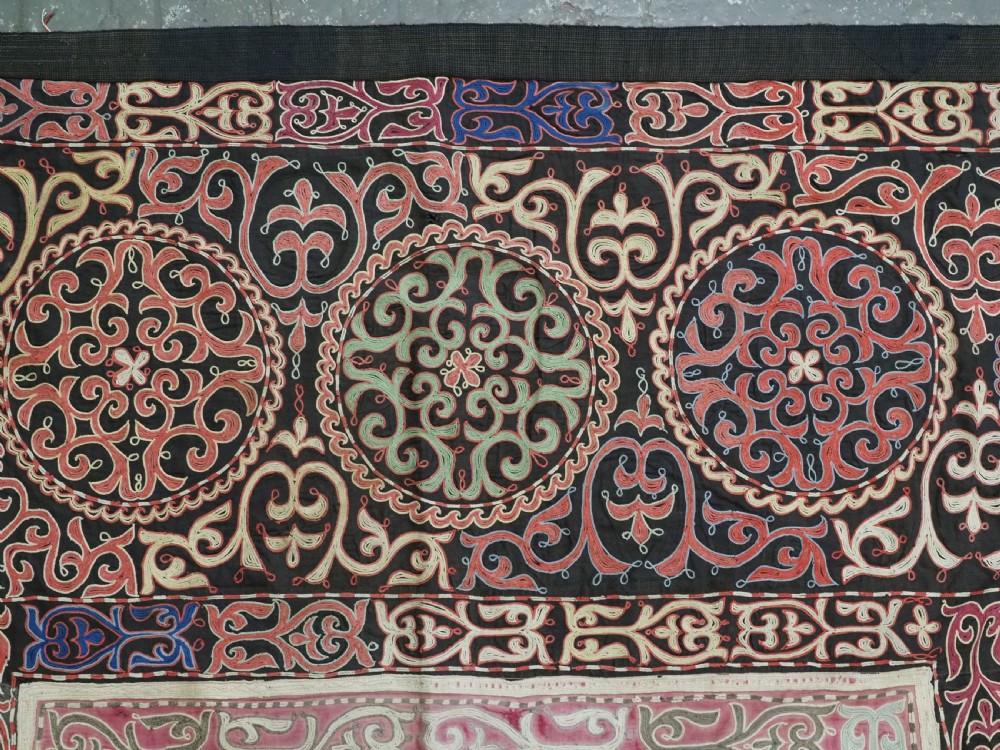 An antique Kyrgyz tush kyiz embroidered wall hanging featuring circular scrollwork medallions and geometric borders in red, pink, and white thread against a dark background. View III
