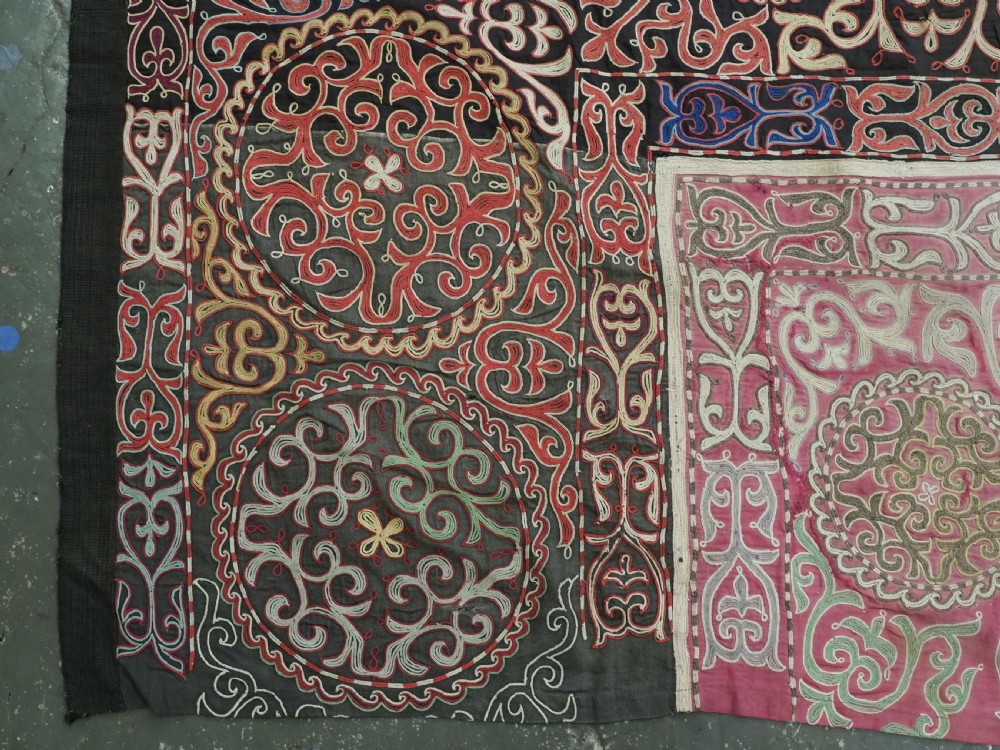 An antique Kyrgyz tush kyiz embroidered wall hanging featuring circular scrollwork medallions and geometric borders in red, pink, and white thread against a dark background. View V