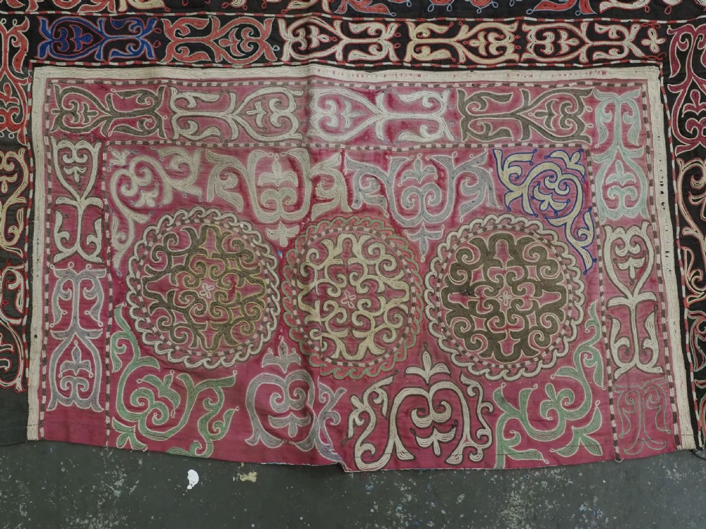 An antique Kyrgyz tush kyiz embroidered wall hanging featuring circular scrollwork medallions and geometric borders in red, pink, and white thread against a dark background. View VI