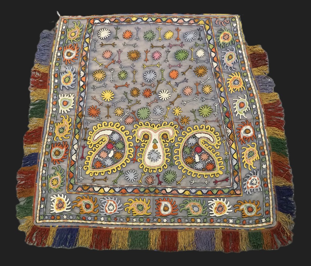 An early 20th-century Uzbek Lakai embroidered saddle cover featuring vibrant silk boteh and floral motifs on a grey ground with colourful fringe trim.