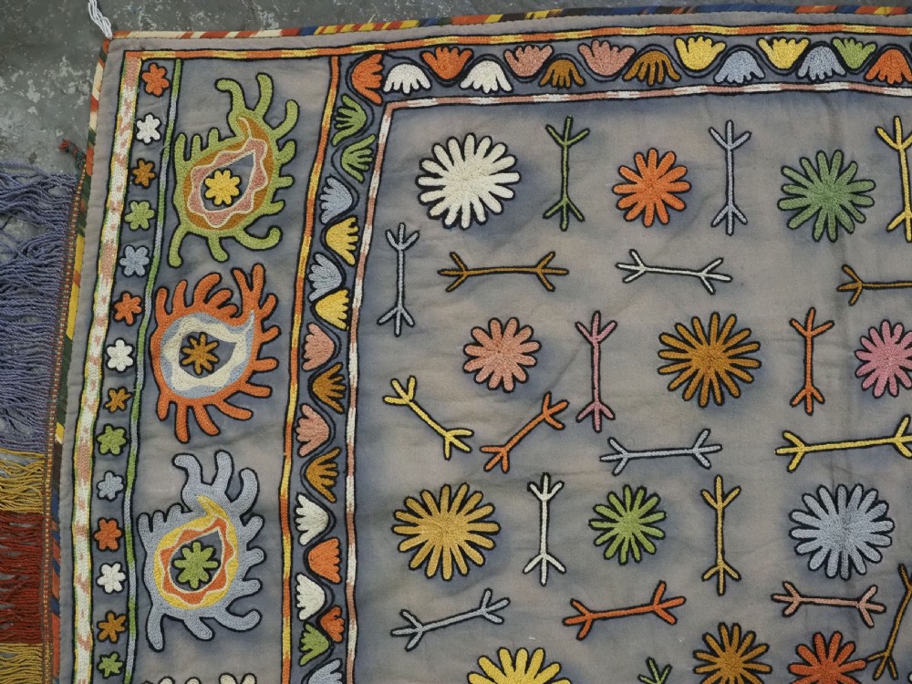 An early 20th-century Uzbek Lakai embroidered saddle cover featuring vibrant silk boteh and floral motifs on a grey ground with colourful fringe trim. View V