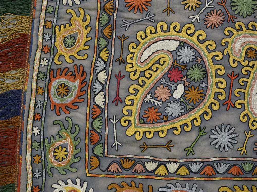 An early 20th-century Uzbek Lakai embroidered saddle cover featuring vibrant silk boteh and floral motifs on a grey ground with colourful fringe trim. View IX
