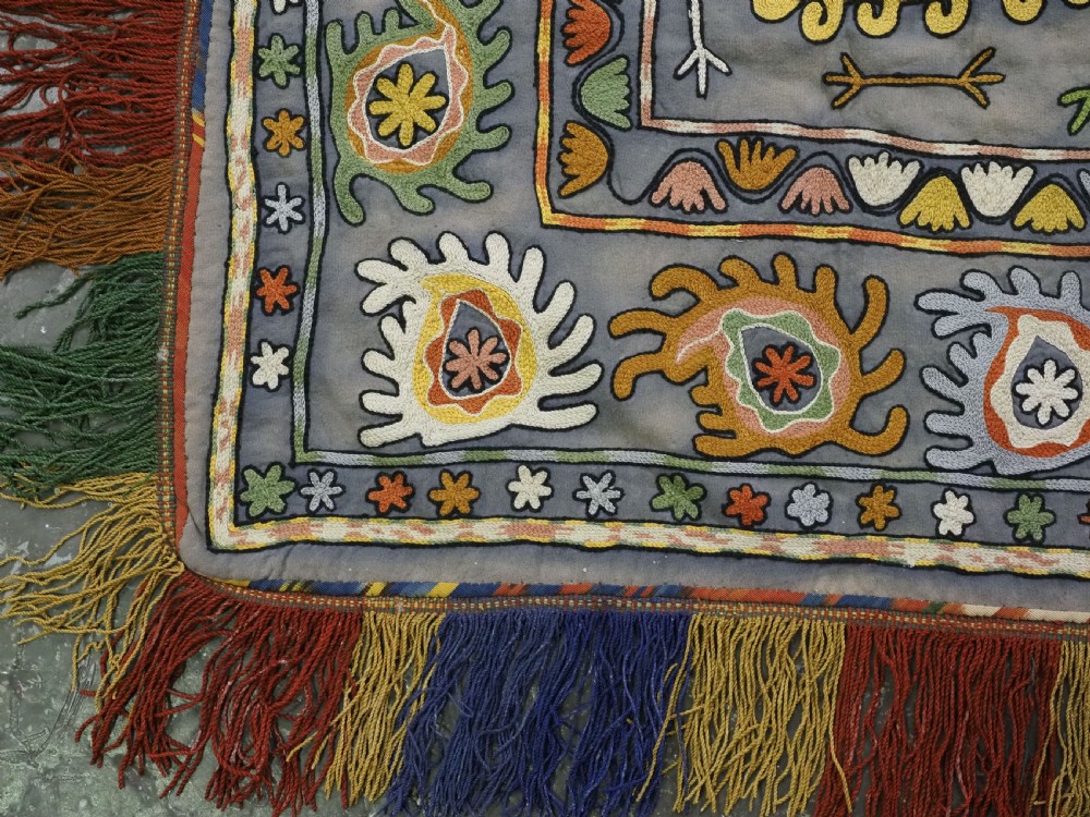 An early 20th-century Uzbek Lakai embroidered saddle cover featuring vibrant silk boteh and floral motifs on a grey ground with colourful fringe trim. View XII