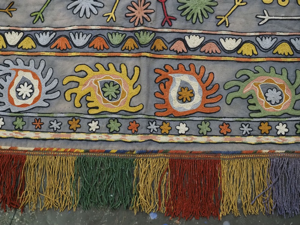 An early 20th-century Uzbek Lakai embroidered saddle cover featuring vibrant silk boteh and floral motifs on a grey ground with colourful fringe trim. View XIII