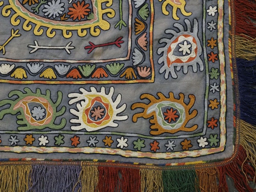 An early 20th-century Uzbek Lakai embroidered saddle cover featuring vibrant silk boteh and floral motifs on a grey ground with colourful fringe trim. View XIV