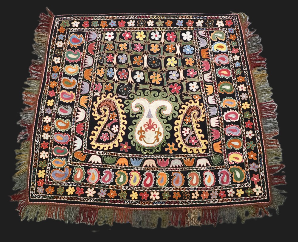 A square black textile decorated with dense multicoloured silk embroidery displaying floral and paisley motifs with fringed edges.