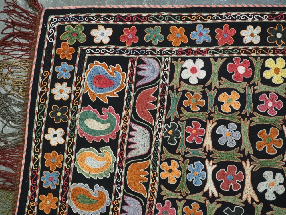 A square black textile decorated with dense multicoloured silk embroidery displaying floral and paisley motifs with fringed edges. View V