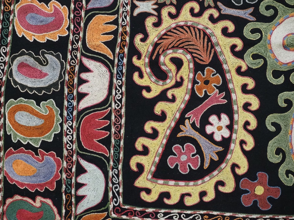 A square black textile decorated with dense multicoloured silk embroidery displaying floral and paisley motifs with fringed edges. View X
