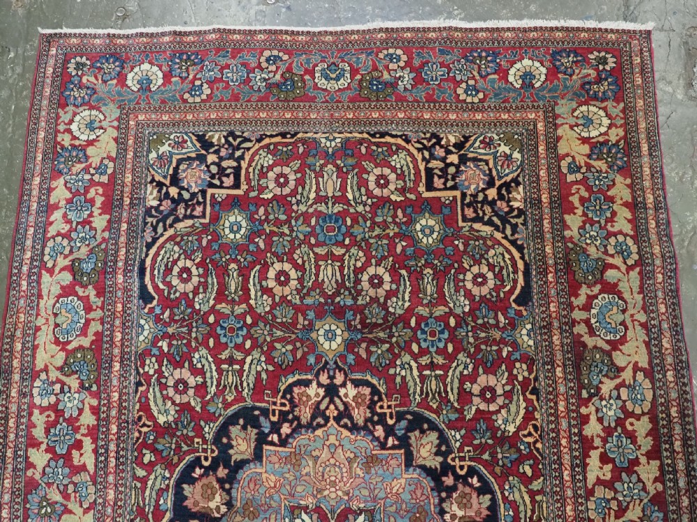 An antique hand-knotted Persian Isfahan wool carpet featuring a central starburst medallion surrounded by dense floral vine motifs in red, navy blue, and cream. View III