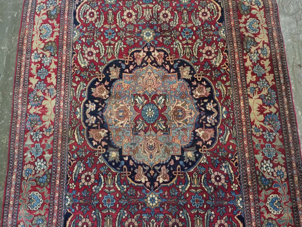 An antique hand-knotted Persian Isfahan wool carpet featuring a central starburst medallion surrounded by dense floral vine motifs in red, navy blue, and cream. View IV