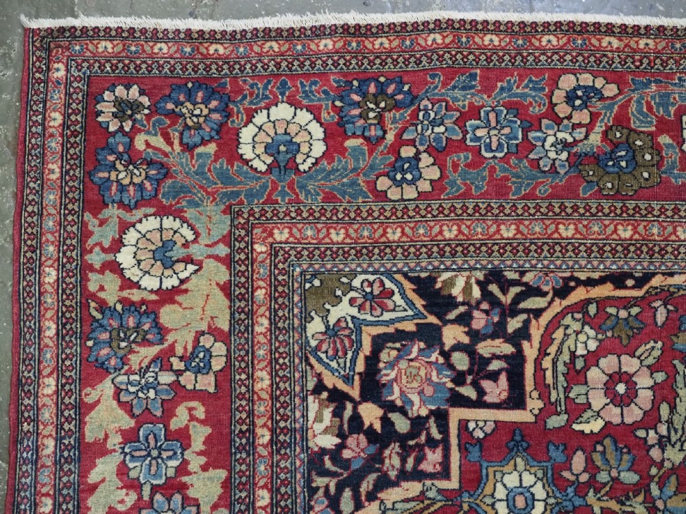 An antique hand-knotted Persian Isfahan wool carpet featuring a central starburst medallion surrounded by dense floral vine motifs in red, navy blue, and cream. View V