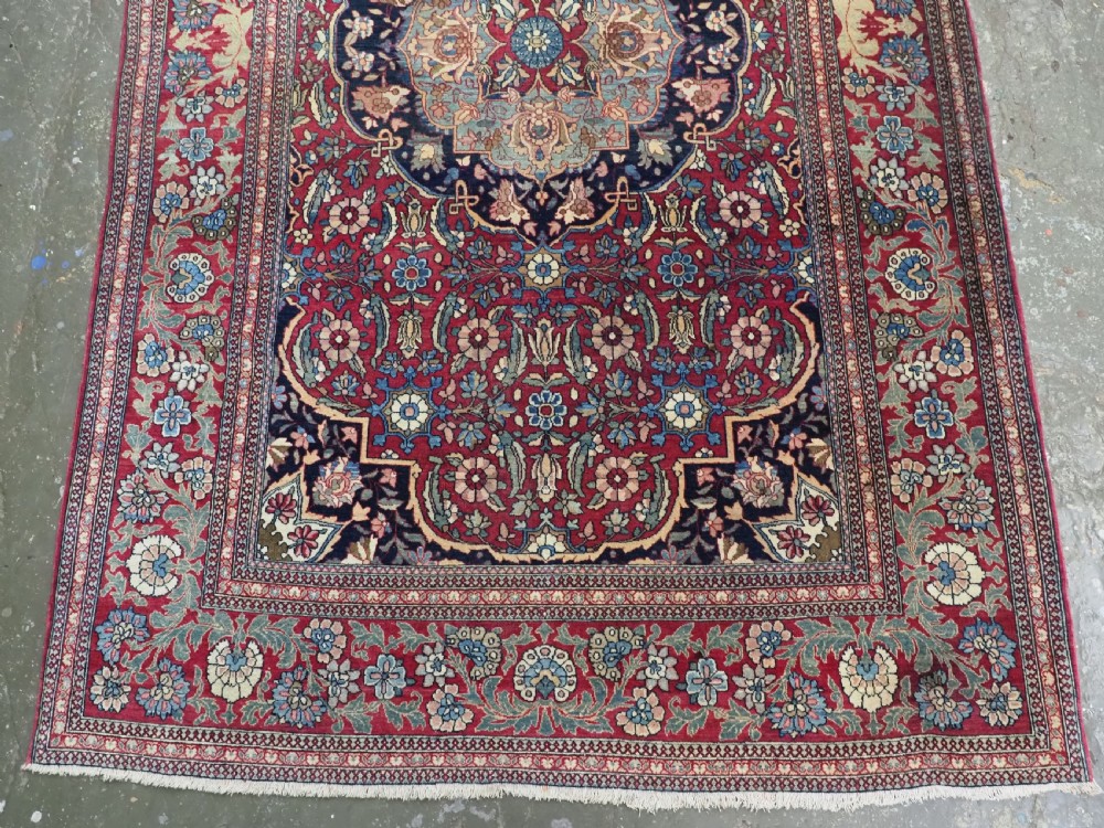 An antique hand-knotted Persian Isfahan wool carpet featuring a central starburst medallion surrounded by dense floral vine motifs in red, navy blue, and cream. View VI