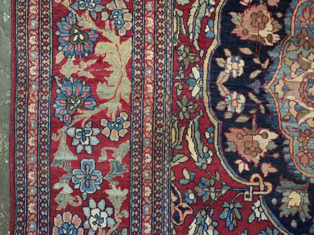 An antique hand-knotted Persian Isfahan wool carpet featuring a central starburst medallion surrounded by dense floral vine motifs in red, navy blue, and cream. View VII