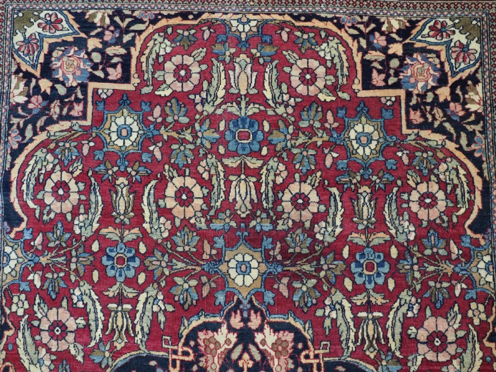 An antique hand-knotted Persian Isfahan wool carpet featuring a central starburst medallion surrounded by dense floral vine motifs in red, navy blue, and cream. View IX