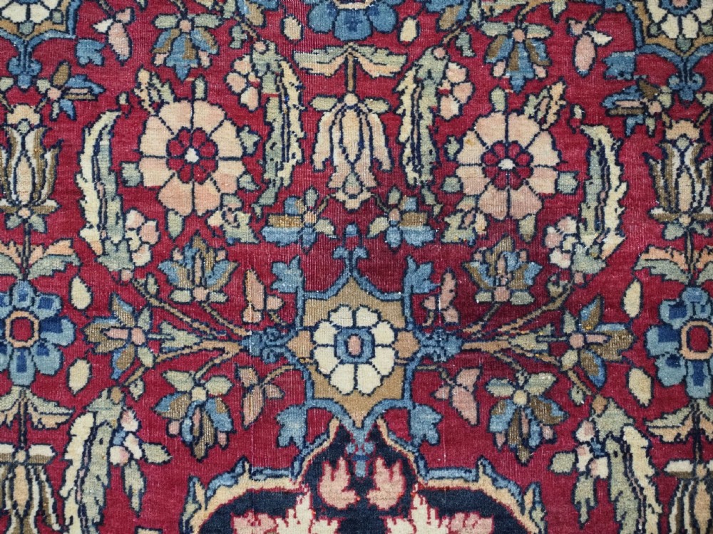 An antique hand-knotted Persian Isfahan wool carpet featuring a central starburst medallion surrounded by dense floral vine motifs in red, navy blue, and cream. View X