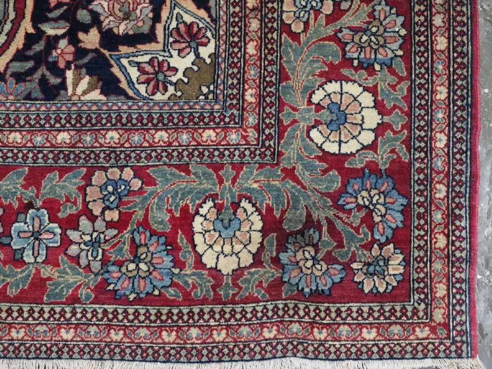 An antique hand-knotted Persian Isfahan wool carpet featuring a central starburst medallion surrounded by dense floral vine motifs in red, navy blue, and cream. View XV