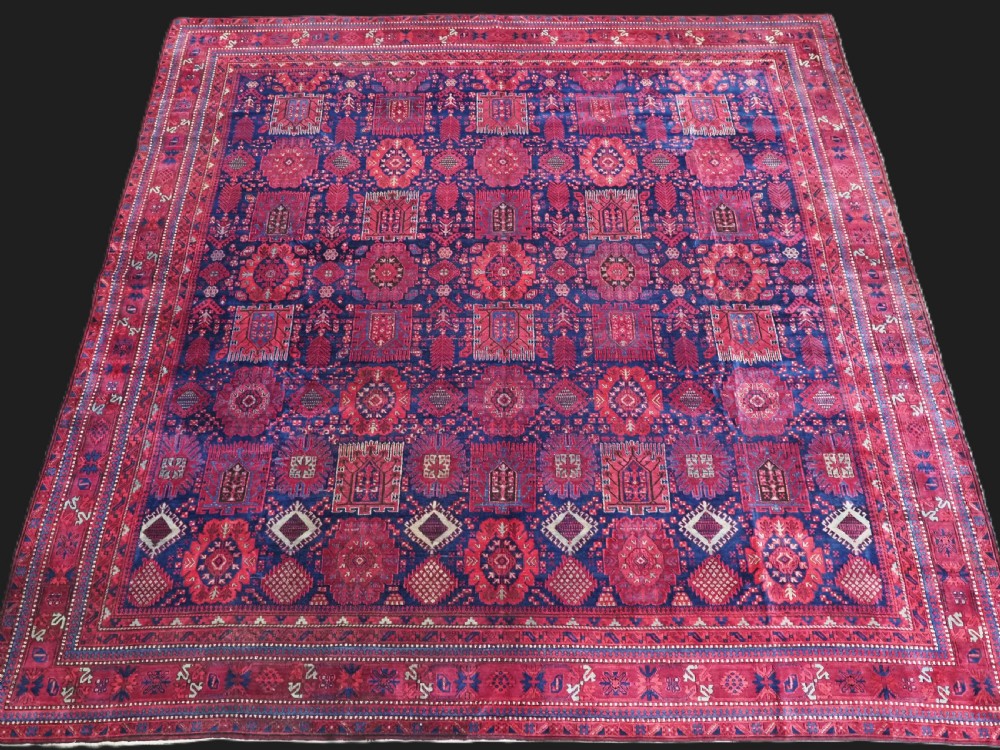 A large hand-knotted wool carpet featuring a repeating geometric medallion pattern in shades of crimson red, deep blue, and magenta.