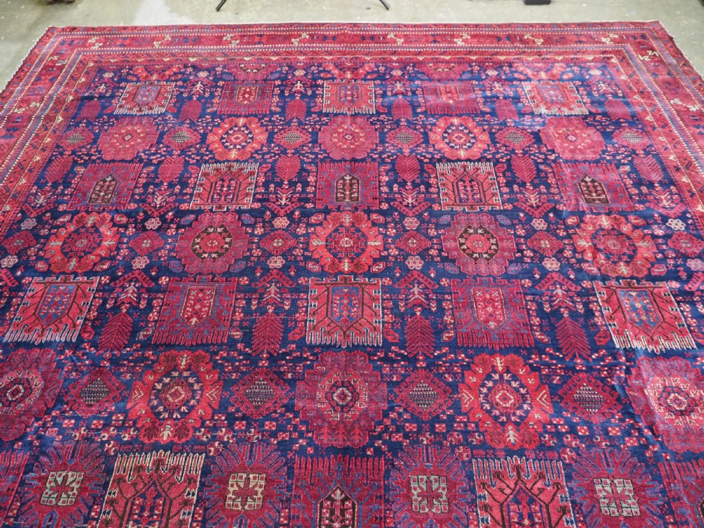 A large hand-knotted wool carpet featuring a repeating geometric medallion pattern in shades of crimson red, deep blue, and magenta. View III