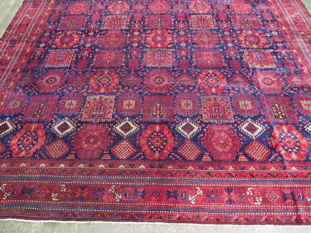 A large hand-knotted wool carpet featuring a repeating geometric medallion pattern in shades of crimson red, deep blue, and magenta. View V