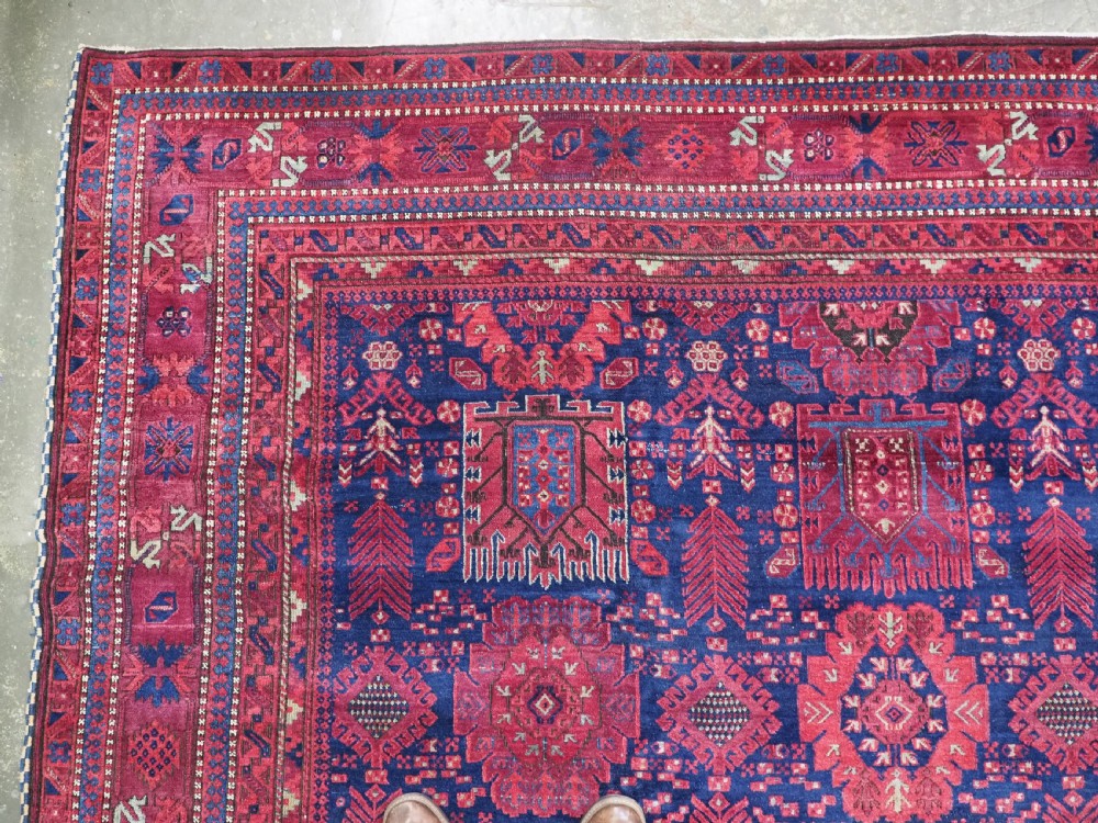 A large hand-knotted wool carpet featuring a repeating geometric medallion pattern in shades of crimson red, deep blue, and magenta. View VI