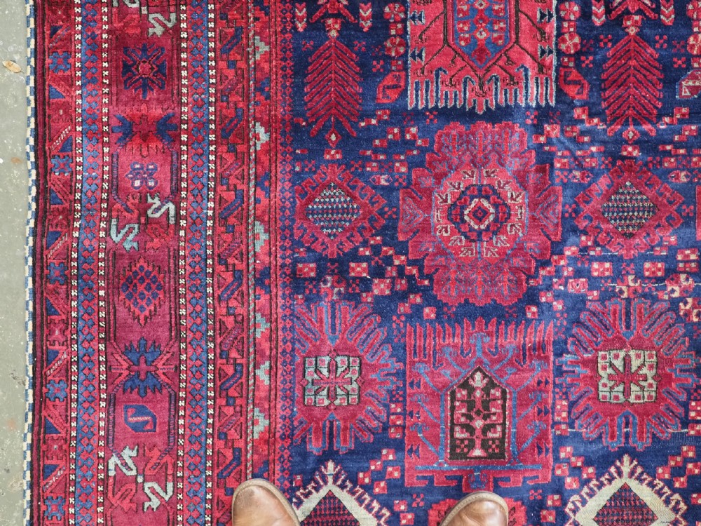 A large hand-knotted wool carpet featuring a repeating geometric medallion pattern in shades of crimson red, deep blue, and magenta. View VII