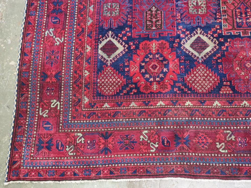 A large hand-knotted wool carpet featuring a repeating geometric medallion pattern in shades of crimson red, deep blue, and magenta. View XII