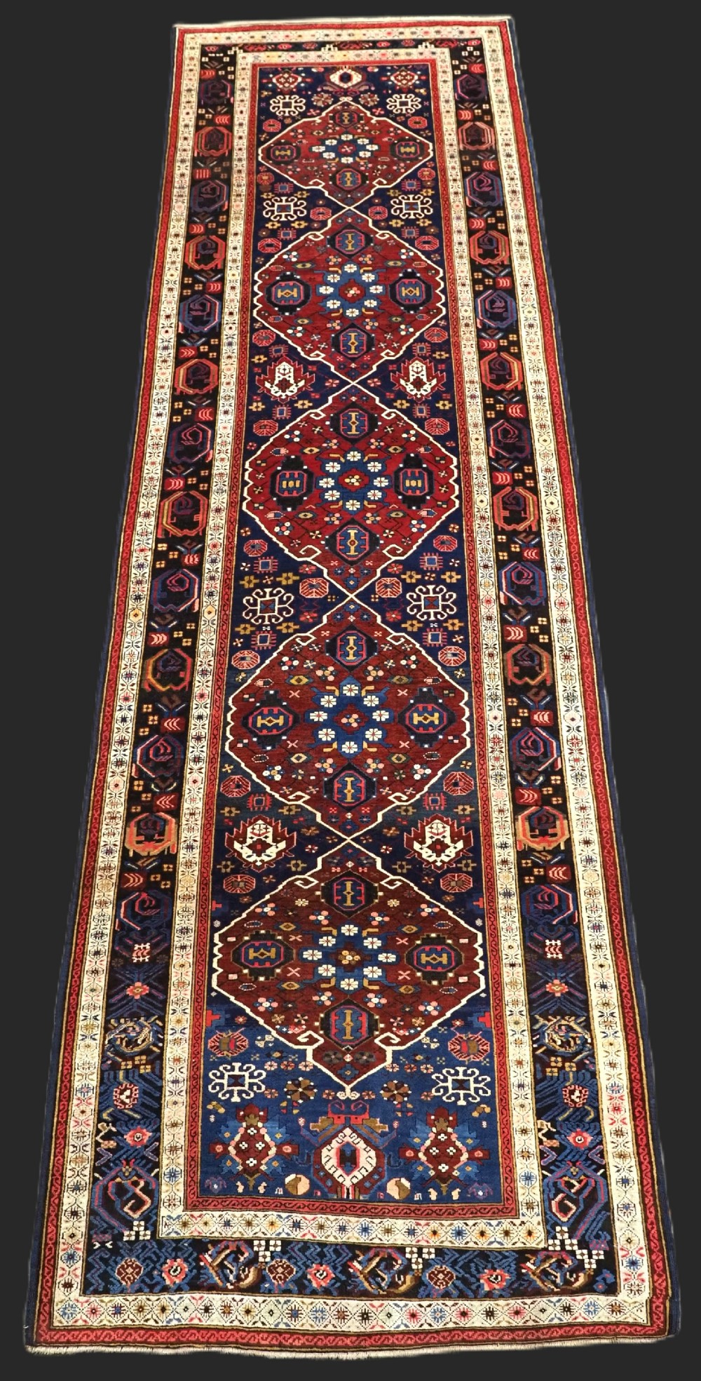 An antique hand-knotted Caucasian Alpan-Kuba runner rug featuring geometric diamond medallions in red and navy blue with detailed ivory borders.