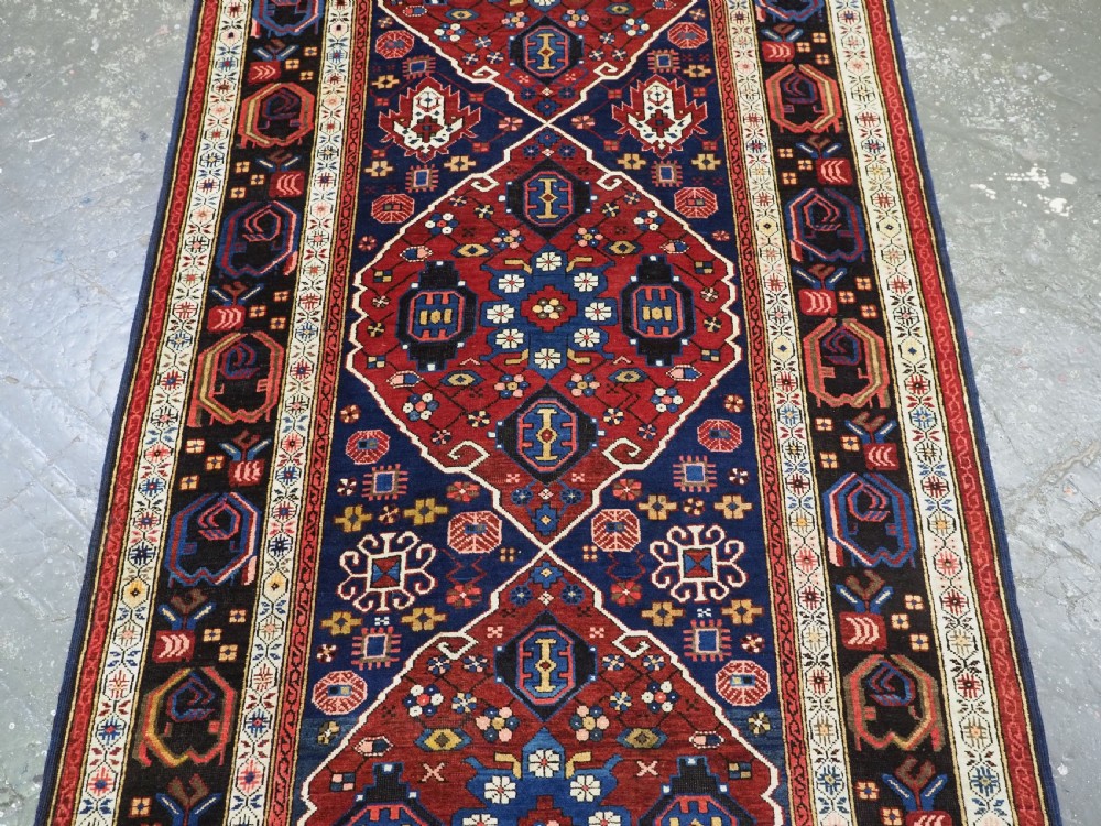 An antique hand-knotted Caucasian Alpan-Kuba runner rug featuring geometric diamond medallions in red and navy blue with detailed ivory borders. View VI