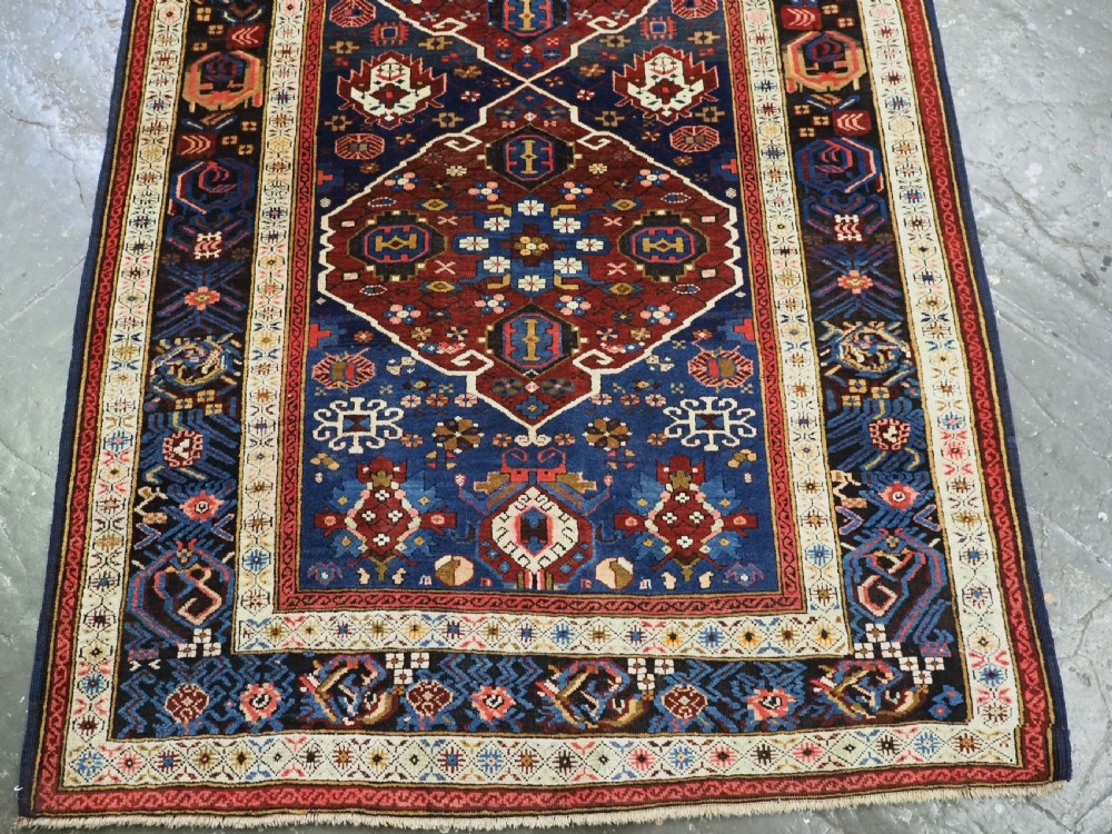 An antique hand-knotted Caucasian Alpan-Kuba runner rug featuring geometric diamond medallions in red and navy blue with detailed ivory borders. View VIII