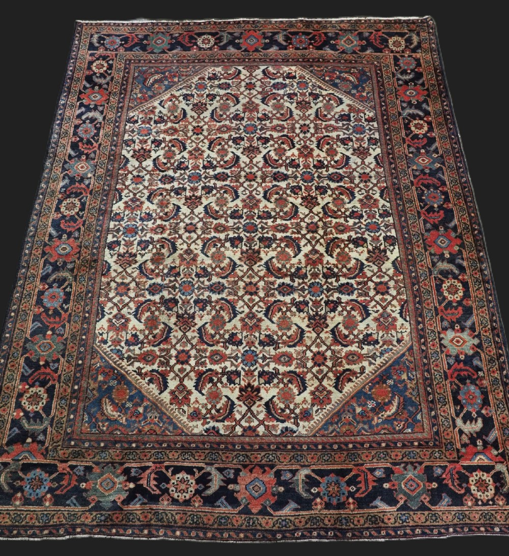 An antique hand-knotted Persian Mahal carpet featuring an ivory central field covered in geometric floral motifs surrounded by dark blue spandrels and a multi-banded border.
