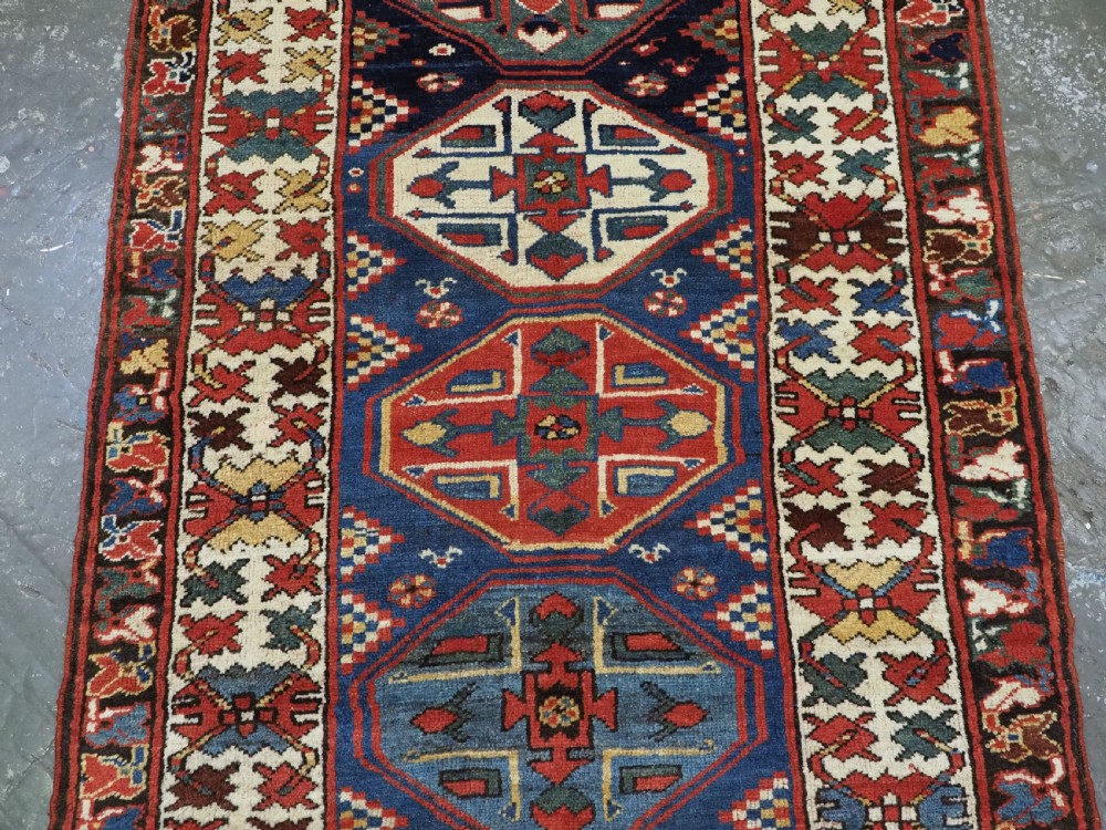 An antique hand-knotted Caucasian wool runner rug featuring a sequence of geometric diamond medallions on an indigo blue field surrounded by patterned borders. View V