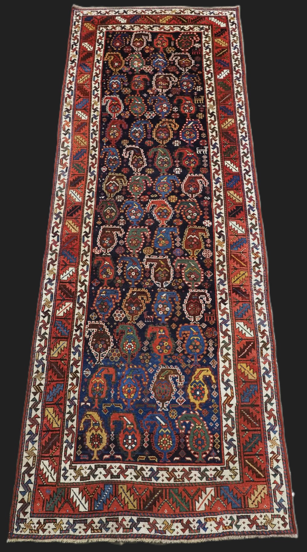 An antique Caucasian Gendje Kazak wool runner rug featuring repeating colourful boteh motifs on a dark navy field framed by geometric red and white borders.