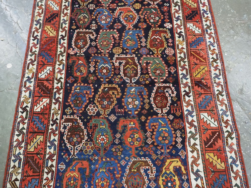 An antique Caucasian Gendje Kazak wool runner rug featuring repeating colourful boteh motifs on a dark navy field framed by geometric red and white borders. View VI
