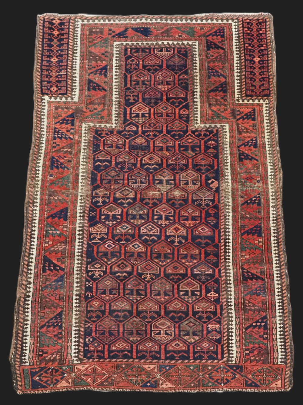 A hand-knotted wool prayer rug with a stepped mihrab niche, repeating geometric tree motifs, and a dark indigo blue and terracotta red palette.