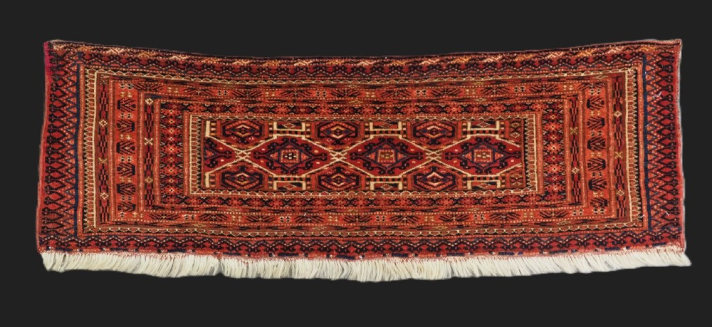 An elongated antique Turkmen torba textile displaying geometric diamond medallion motifs in deep red, dark navy, and cream wool with fringed bottom edge.