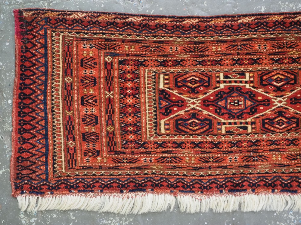 An elongated antique Turkmen torba textile displaying geometric diamond medallion motifs in deep red, dark navy, and cream wool with fringed bottom edge. View II