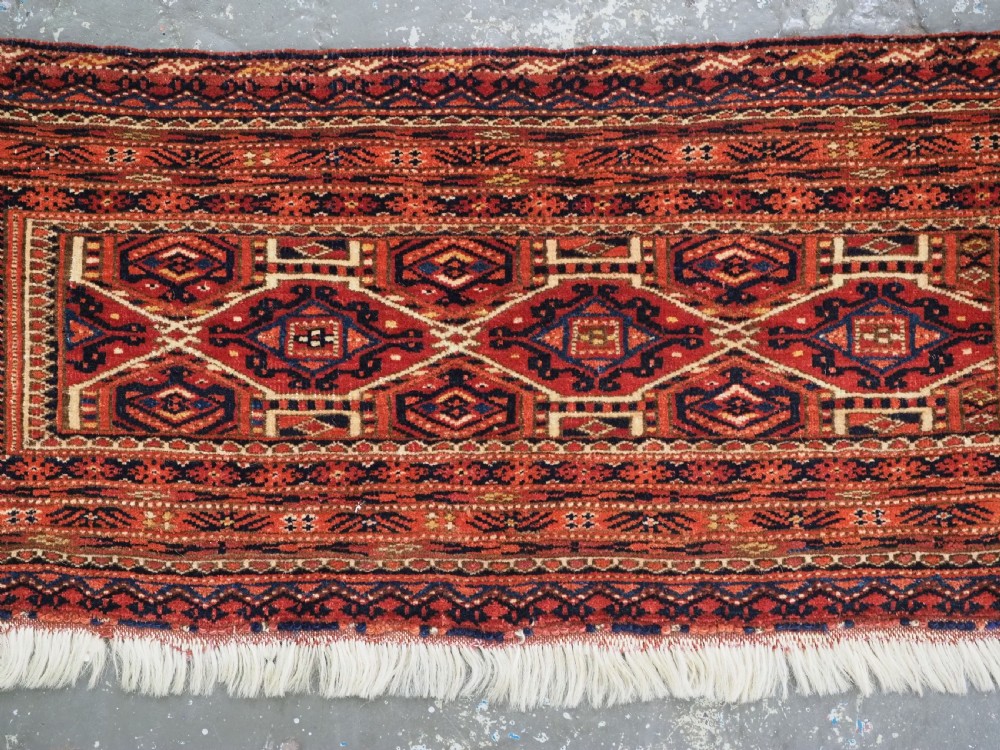 An elongated antique Turkmen torba textile displaying geometric diamond medallion motifs in deep red, dark navy, and cream wool with fringed bottom edge. View III